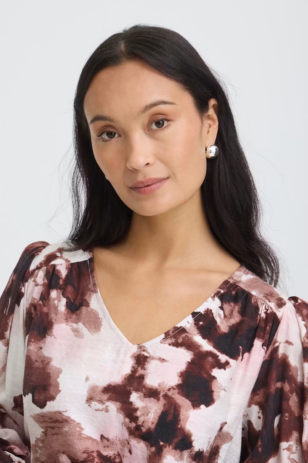 A woman with long dark hair wearing a Watercolour Bloom Rum Raisin / Red V-neck blouse photographed against a light gray studio background.