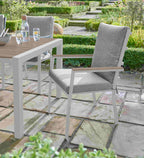 White outdoor dining chair with a light wood armrest and a gray textured cushion, shown on a stone patio next to a wooden table in a landscaped garden.