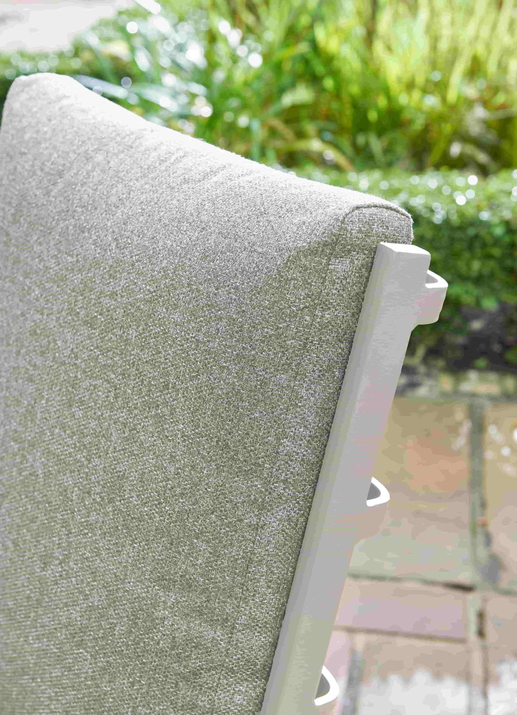 Close-up of a light gray textured outdoor patio cushion on a white-framed chair, with a blurred garden and stone patio background.