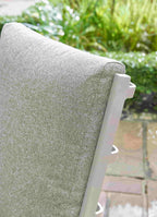 Close-up of a light gray textured outdoor patio cushion on a white-framed chair, with a blurred garden and stone patio background.
