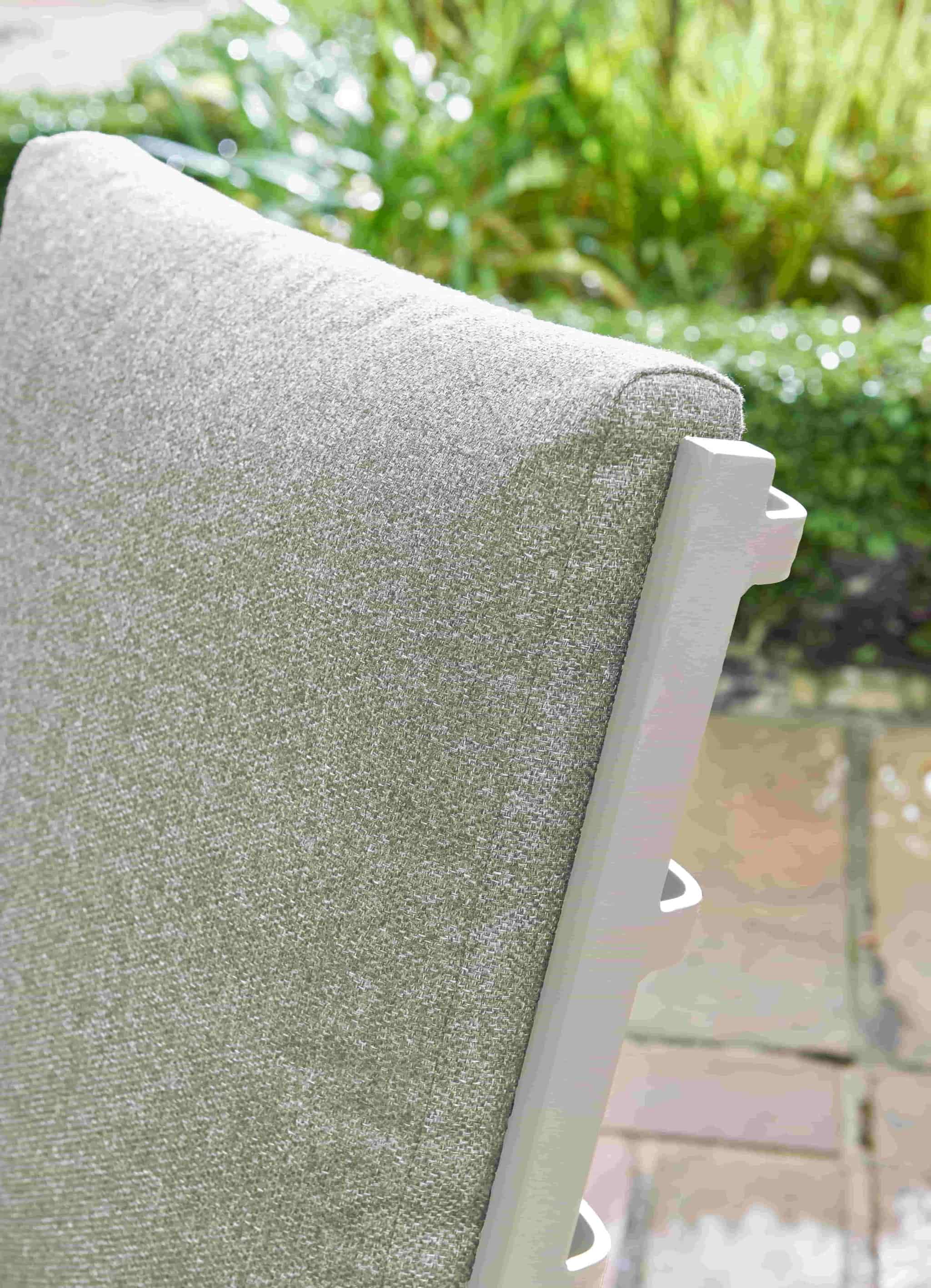 Close-up of a light gray textured outdoor patio cushion on a white-framed chair, with a blurred garden and stone patio background.