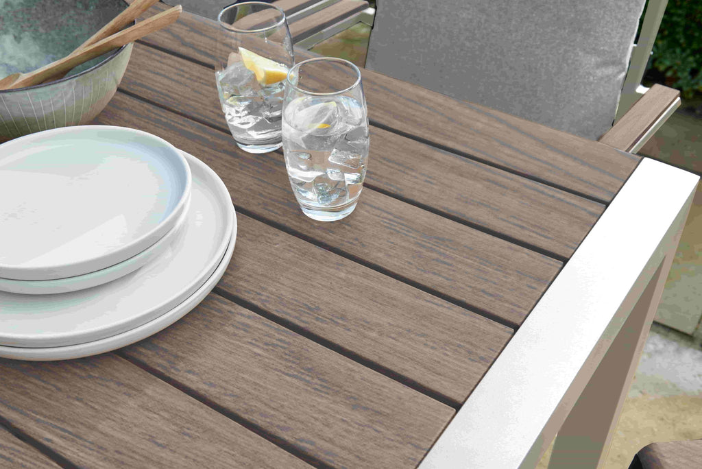 Outdoor dining table with brown slatted surfaces, white plates, and two clear drinking glasses with water and a lemon slice, shown in a real patio setting.