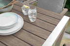 Outdoor dining table with brown slatted surfaces, white plates, and two clear drinking glasses with water and a lemon slice, shown in a real patio setting.