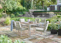 An outdoor dining set consisting of a rectangular table and six matching chairs, placed on a stone patio in a landscaped garden with trees, plants, and a stone wall in the background.