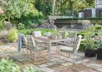 An outdoor dining set consisting of a rectangular table and six matching chairs, placed on a stone patio in a landscaped garden with trees, plants, and a stone wall in the background.