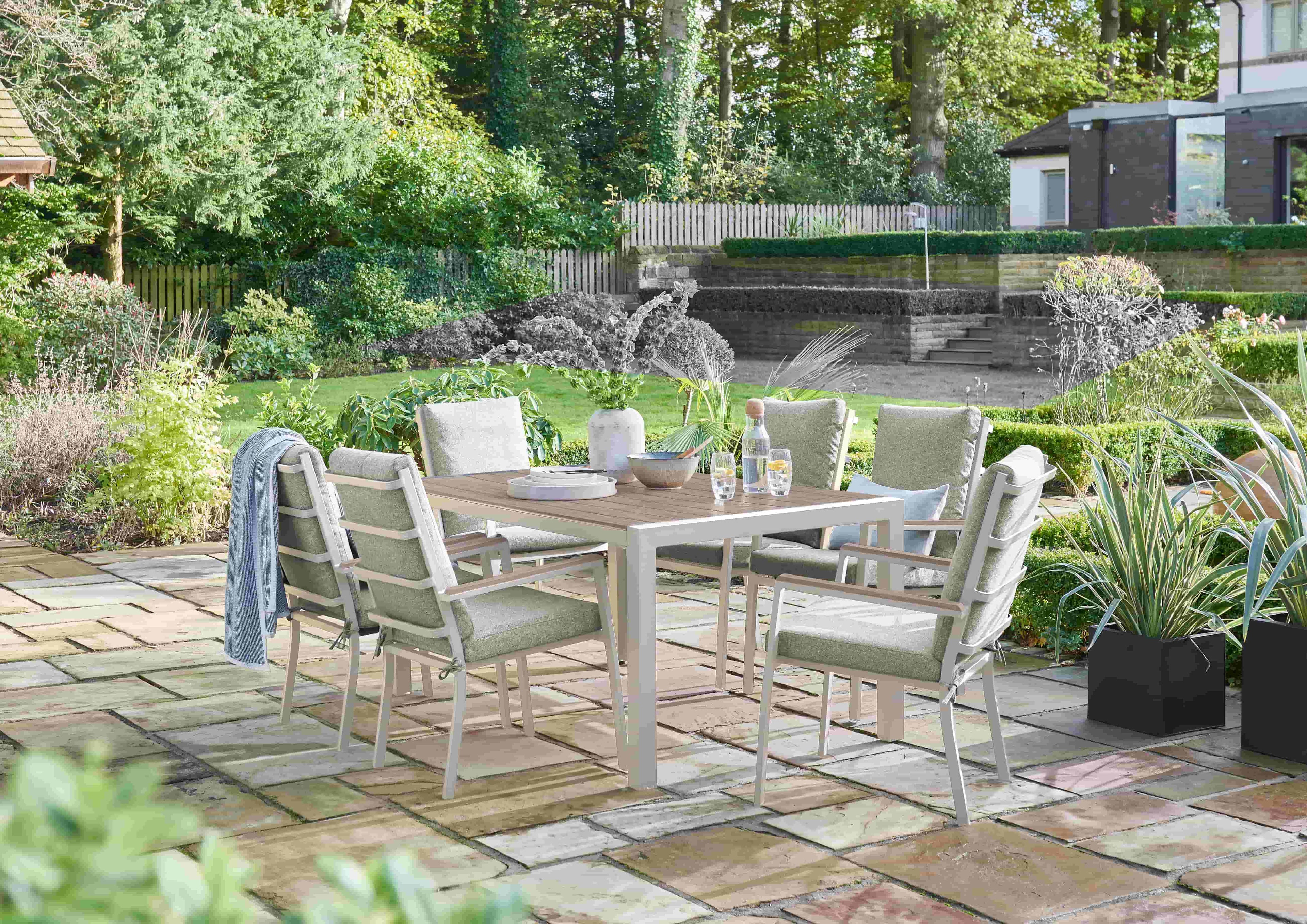 An outdoor dining set consisting of a rectangular table and six matching chairs, placed on a stone patio in a landscaped garden with trees, plants, and a stone wall in the background.