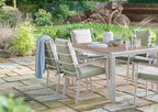 Outdoor dining set on a stone patio in a garden, featuring a rectangular wooden table with a light gray frame and six matching armchairs with light green cushions and a blue throw blanket.