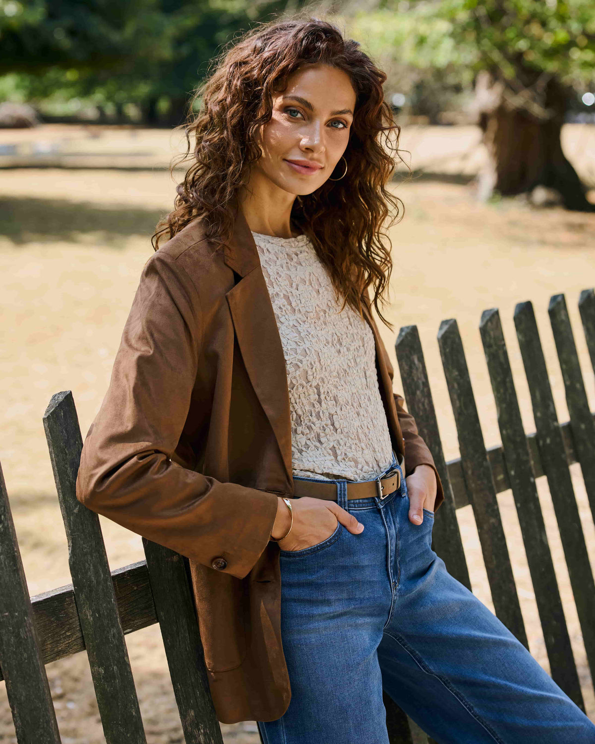 A woman wearing a brown blazer, cream lace top, and blue jeans leans against a dark wooden fence in an outdoor park setting.