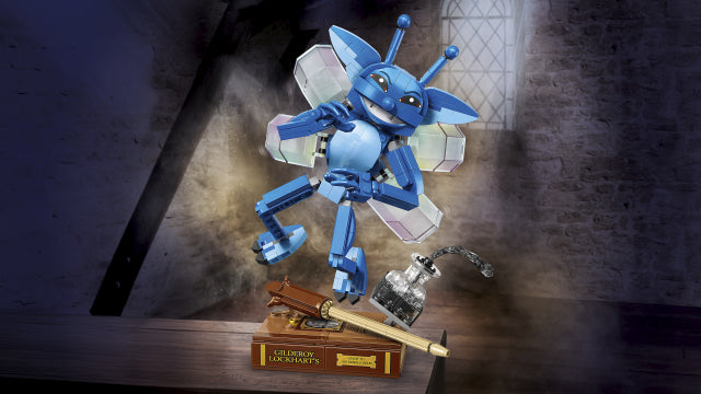 A blue robotic insect figure with translucent wings stands on a wooden surface in a dimly lit room, with a small book, a wooden rod, and a glass bottle nearby.