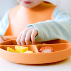 Child's hand reaching for food on an orange plate with a bib