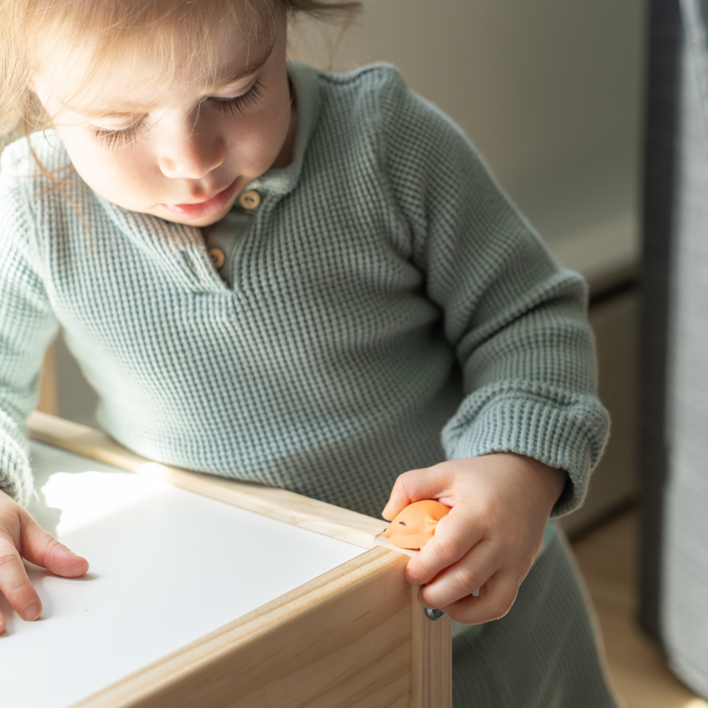 Child in a light green sweater holding a small toy, sitting at a wooden table.
