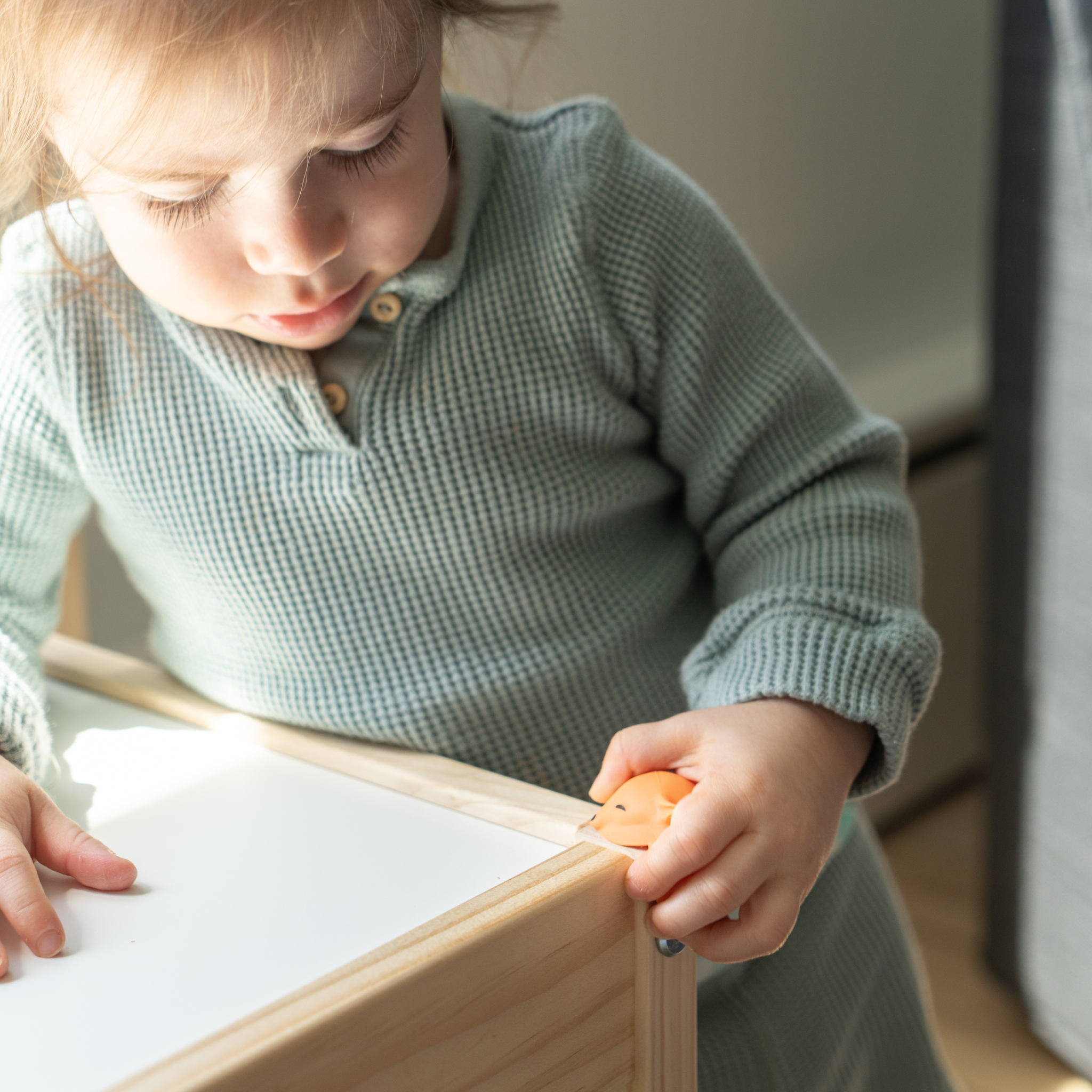 Child in a light green sweater holding a small toy, sitting at a wooden table.