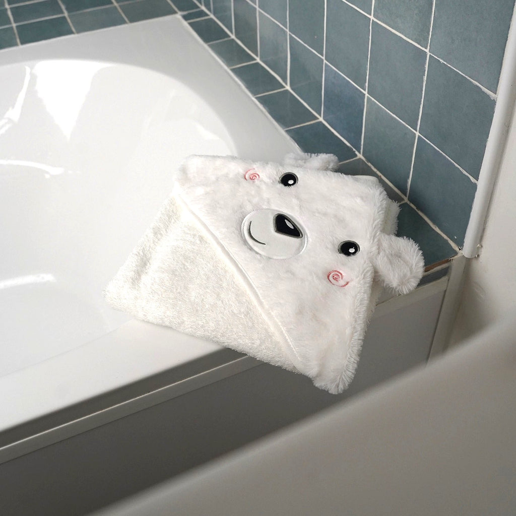 A white plush bath towel shaped like a bear face with embroidered black eyes, nose, and pink cheeks, draped over the edge of a white bathtub in a tiled bathroom.