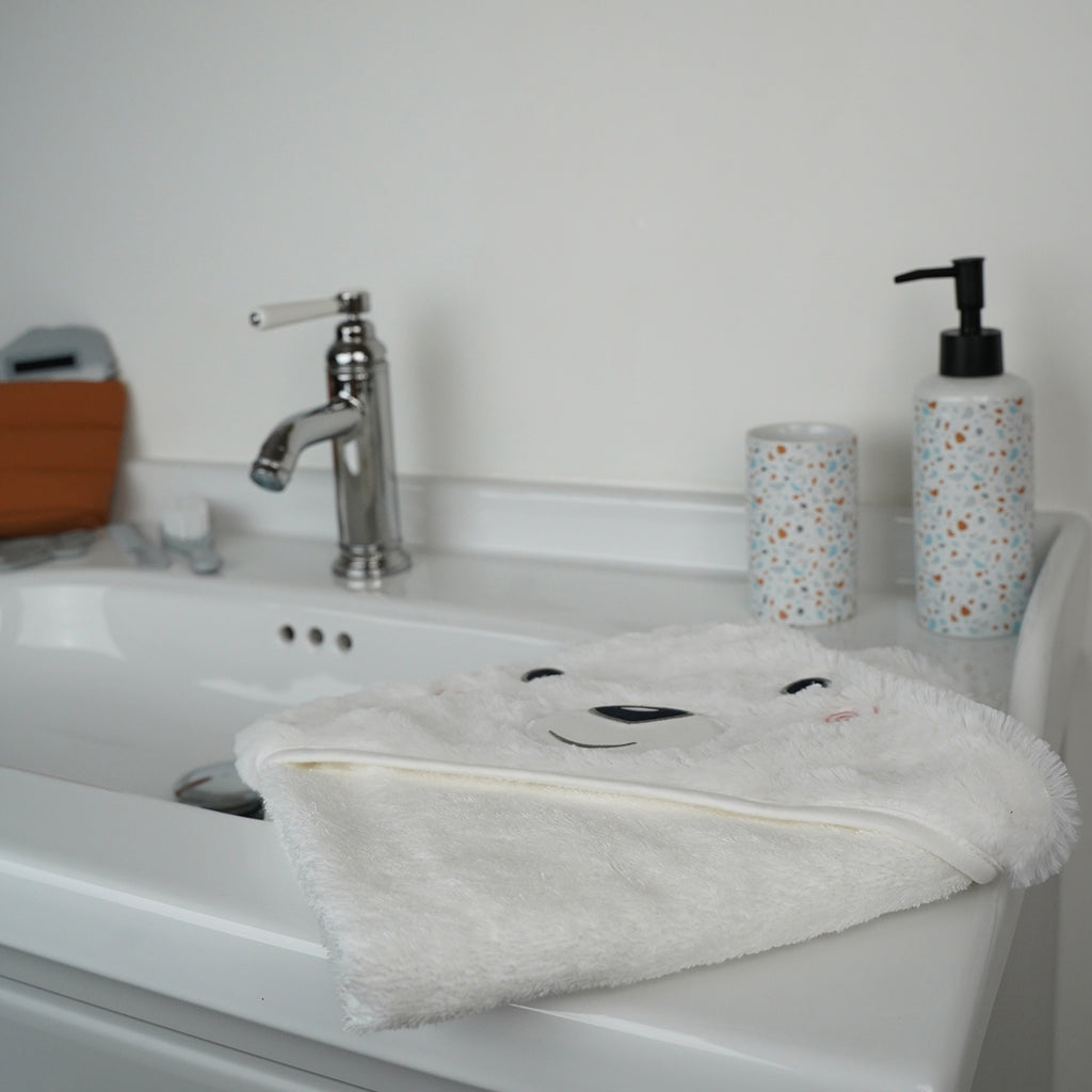 A white plush baby towel with a bear face hood is draped over a white ceramic bathroom sink, with a chrome faucet and patterned soap dispenser in the background.