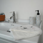 A white plush baby towel with a bear face hood is draped over a white ceramic bathroom sink, with a chrome faucet and patterned soap dispenser in the background.