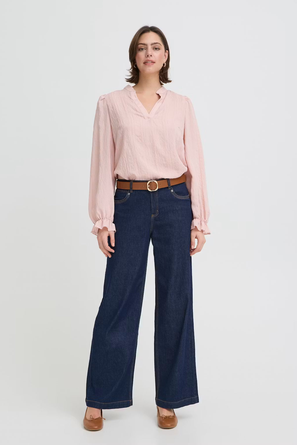 A woman modeling a light pink long-sleeve blouse with ruffled cuffs, paired with dark blue wide-leg jeans, a brown belt, and brown shoes, against a plain light gray studio background.