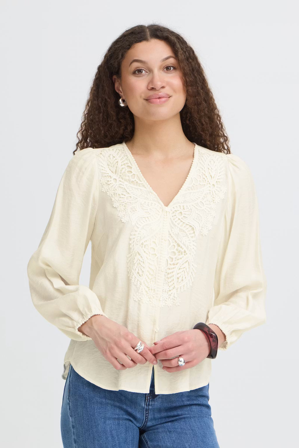 A woman with curly brown hair wearing a Whisper White long-sleeve blouse with lace detailing on the V-neckline, paired with blue jeans, against a light gray studio background.