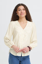 A woman with curly brown hair wearing a Whisper White long-sleeve blouse with lace detailing on the V-neckline, paired with blue jeans, against a light gray studio background.
