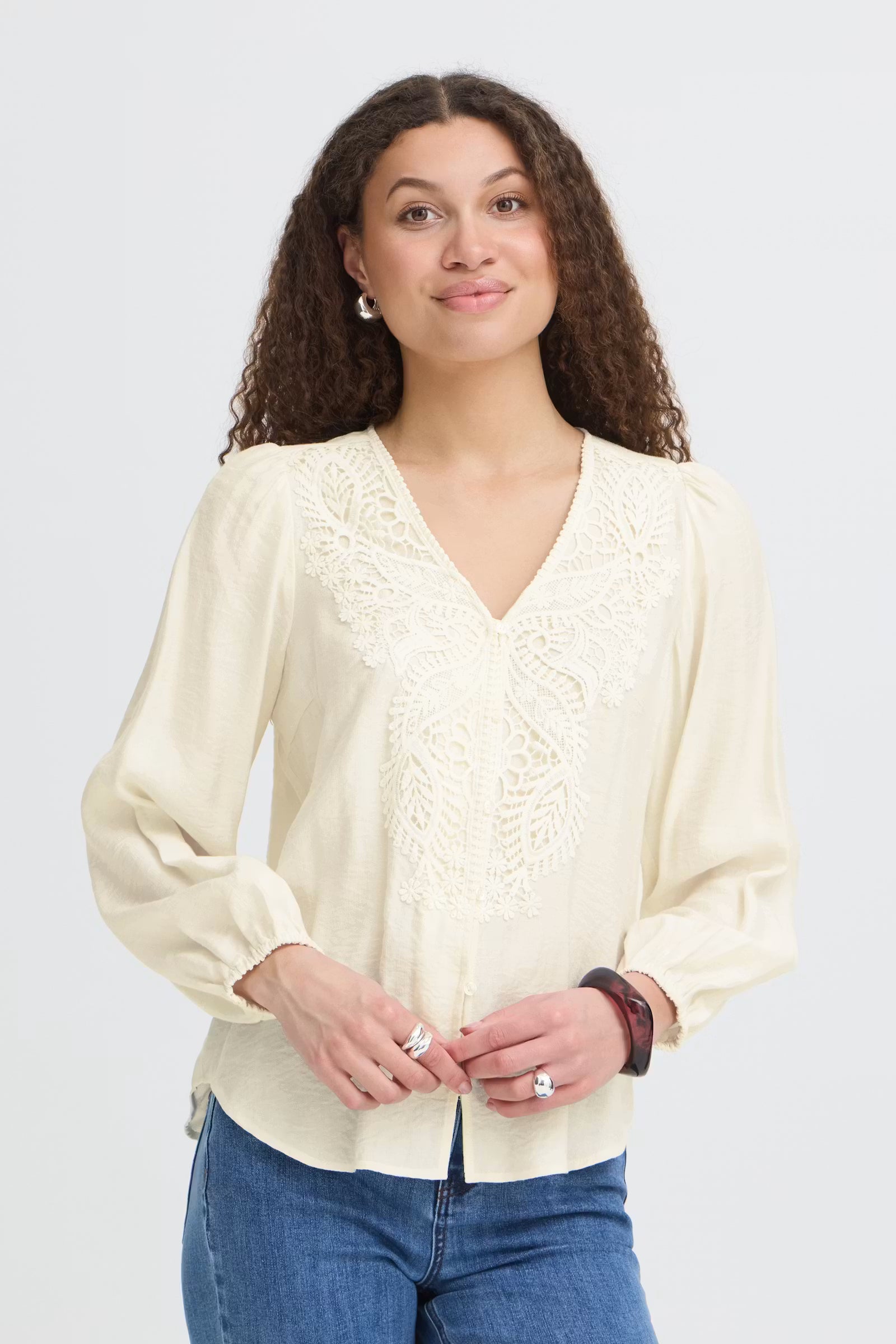 A woman with curly brown hair wearing a Whisper White long-sleeve blouse with lace detailing on the V-neckline, paired with blue jeans, against a light gray studio background.