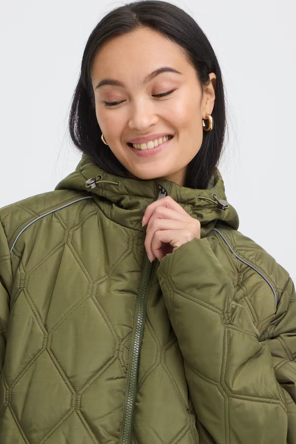 A woman wearing FRAGATE Outerwear in Grape Leaf/Green quilted puffer jacket with a high collar and zipper, shown in a close-up shot against a light gray background.
