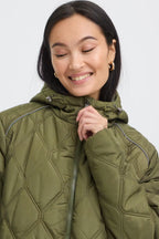A woman wearing FRAGATE Outerwear in Grape Leaf/Green quilted puffer jacket with a high collar and zipper, shown in a close-up shot against a light gray background.