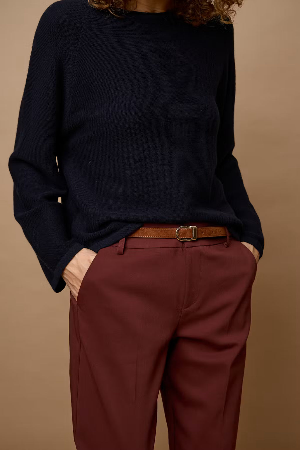 A person wearing a dark navy knit sweater and 
Rum Raisin / Red trousers with a brown belt, shown from the neck down against a plain warm beige studio background.