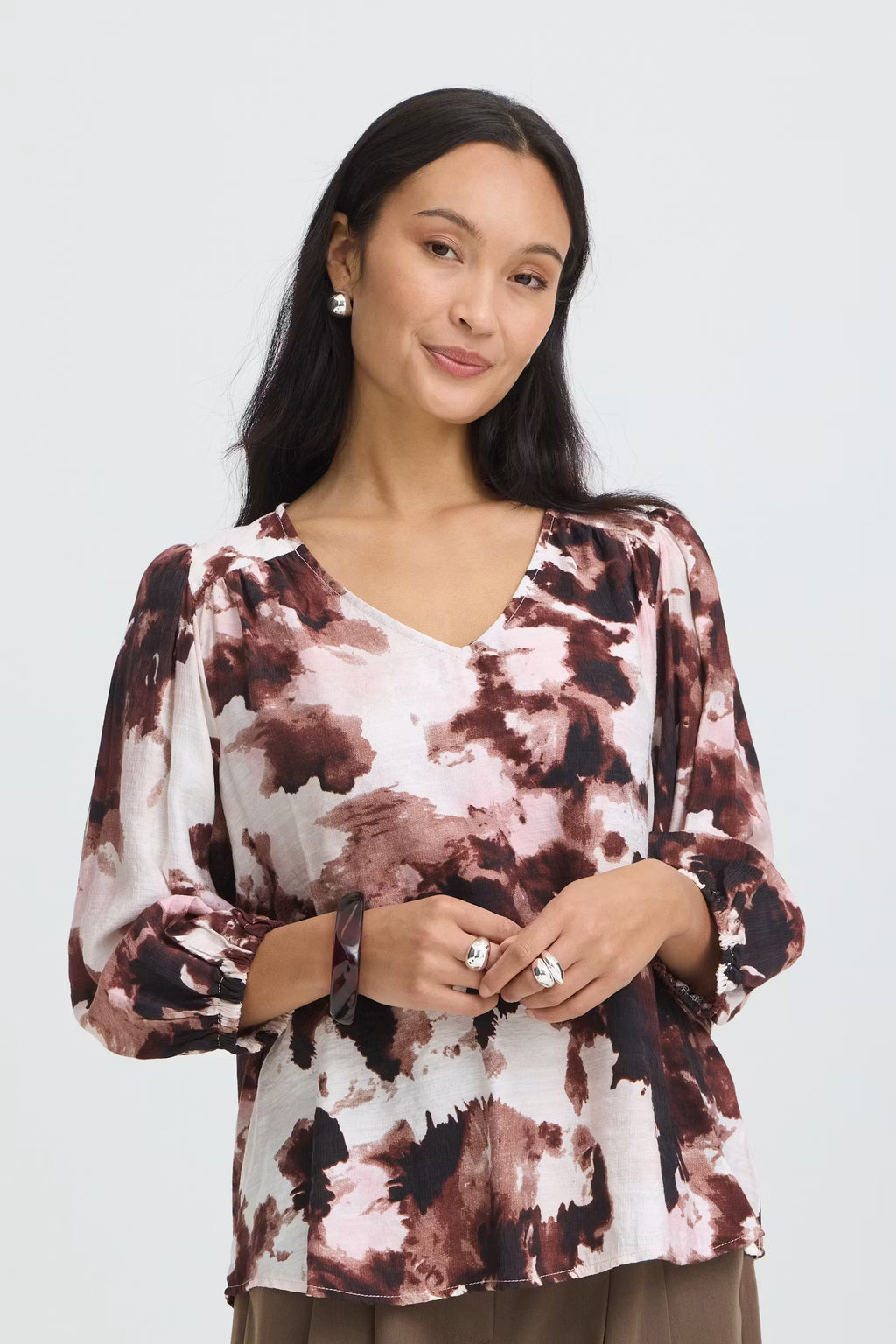 A woman wearing a long-sleeve Watercolour Bloom Rum Raisin / Red V-neck blouse photographed against a light gray studio background.