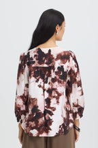 A woman viewed from the back wearing a Watercolour Bloom Rum Raisin / Red long-sleeve blouse  paired with brown pants, against a light gray studio background.