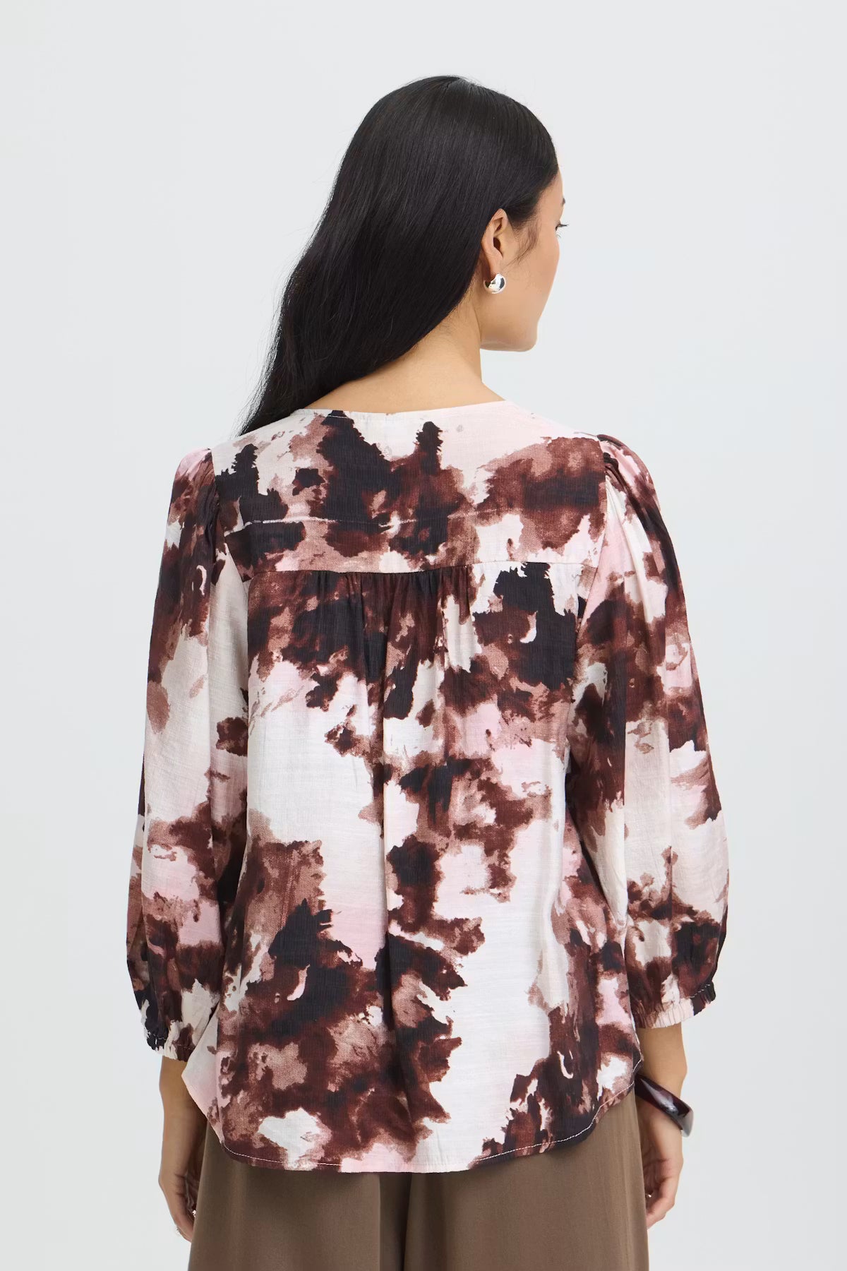 A woman viewed from the back wearing a Watercolour Bloom Rum Raisin / Red long-sleeve blouse  paired with brown pants, against a light gray studio background.