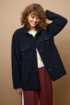 A woman wearing a dark navy textured bouclé jacket over a cream blouse and burgundy trousers, standing against a solid tan studio background.