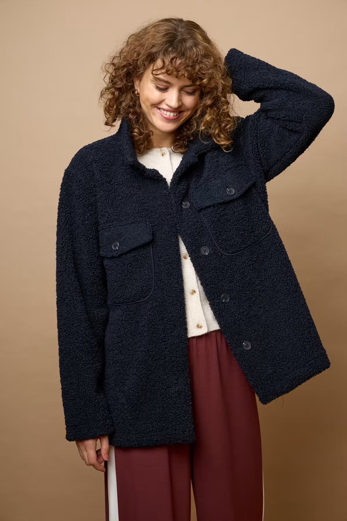 A woman wearing a dark navy textured bouclé jacket over a cream blouse and burgundy trousers, standing against a solid tan studio background.