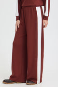 A person wearing wide-leg burgundy sweatpants with a bold white vertical stripe down the side, paired with a matching burgundy top and black shoes, shown against a plain light gray studio background.