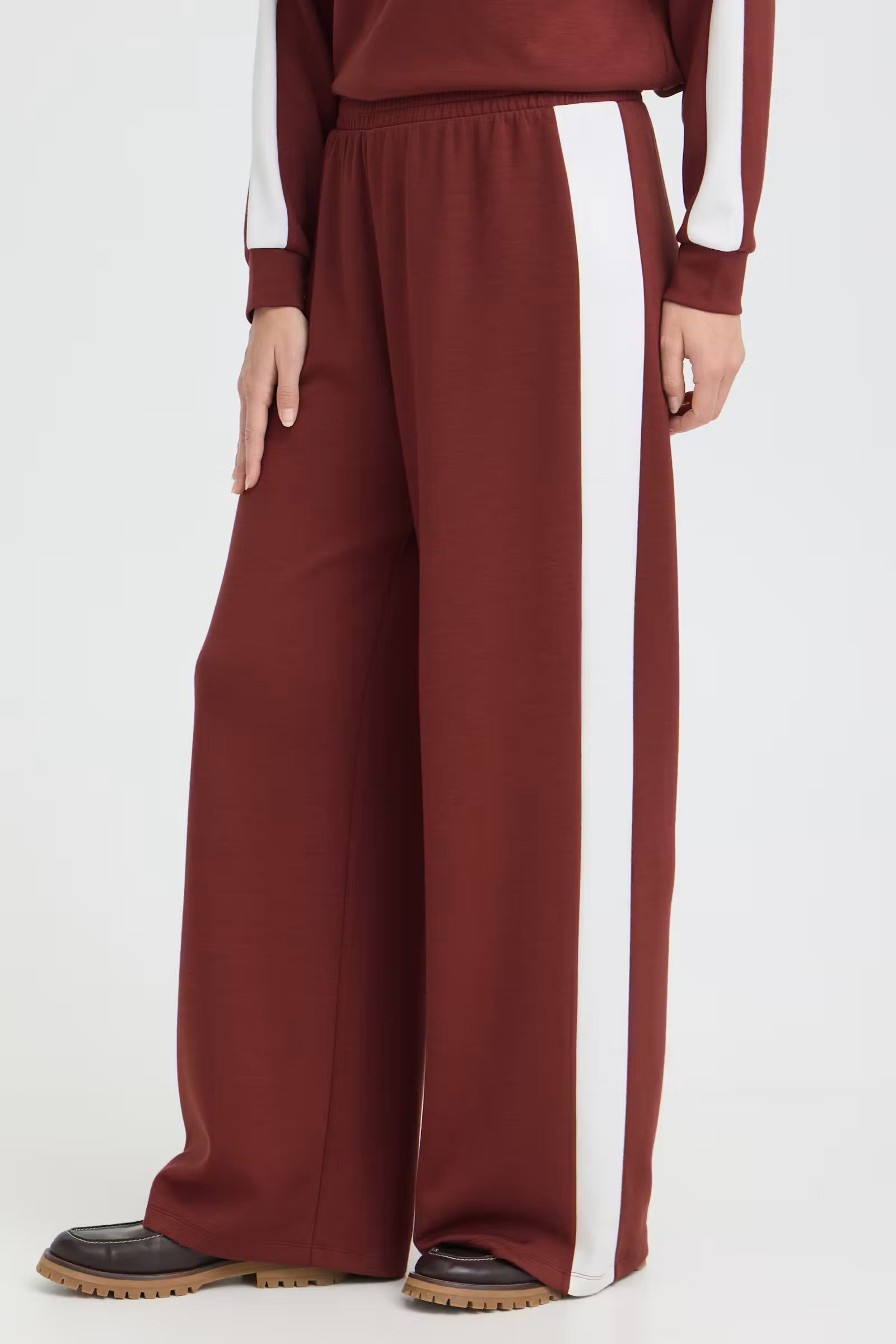 A person wearing wide-leg burgundy sweatpants with a bold white vertical stripe down the side, paired with a matching burgundy top and black shoes, shown against a plain light gray studio background.