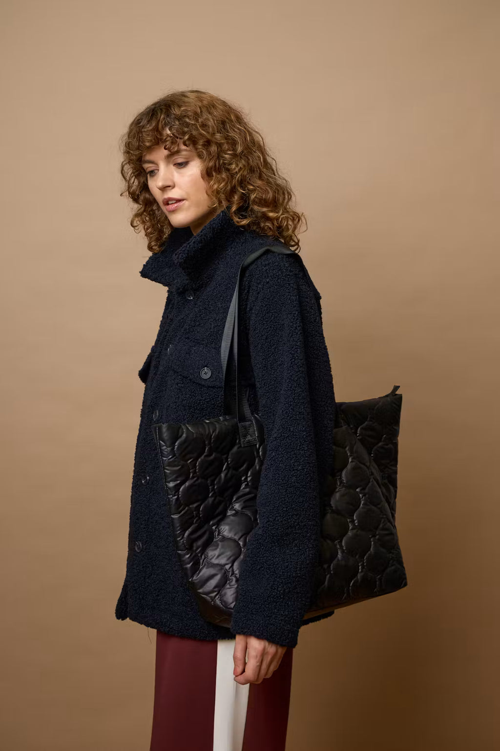 A model with curly brown hair is shown from the side, wearing a dark navy blue textured coat and carrying a large black quilted leather tote bag against a solid tan studio background.