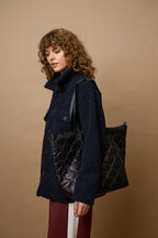A model with curly brown hair is shown from the side, wearing a dark navy blue textured coat and carrying a large black quilted leather tote bag against a solid tan studio background.