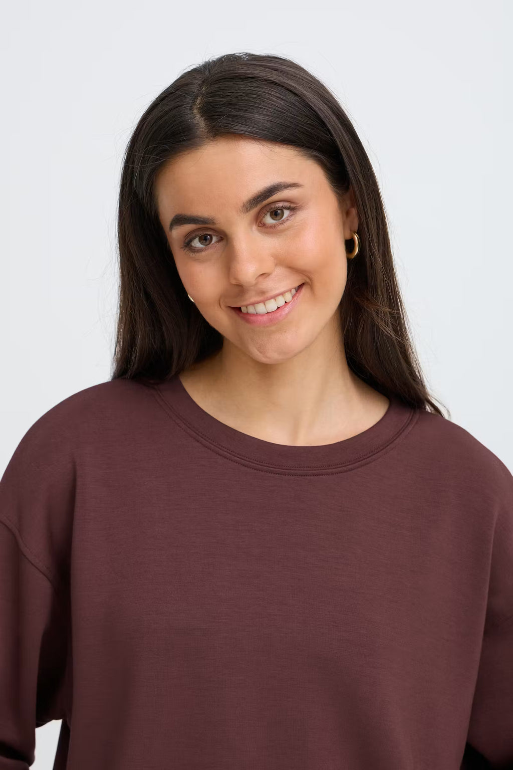 A woman with long dark brown hair wearing a FRAURORA PU 7 Sweatshirt in Rum Raisin/Red , photographed against a light gray studio background.