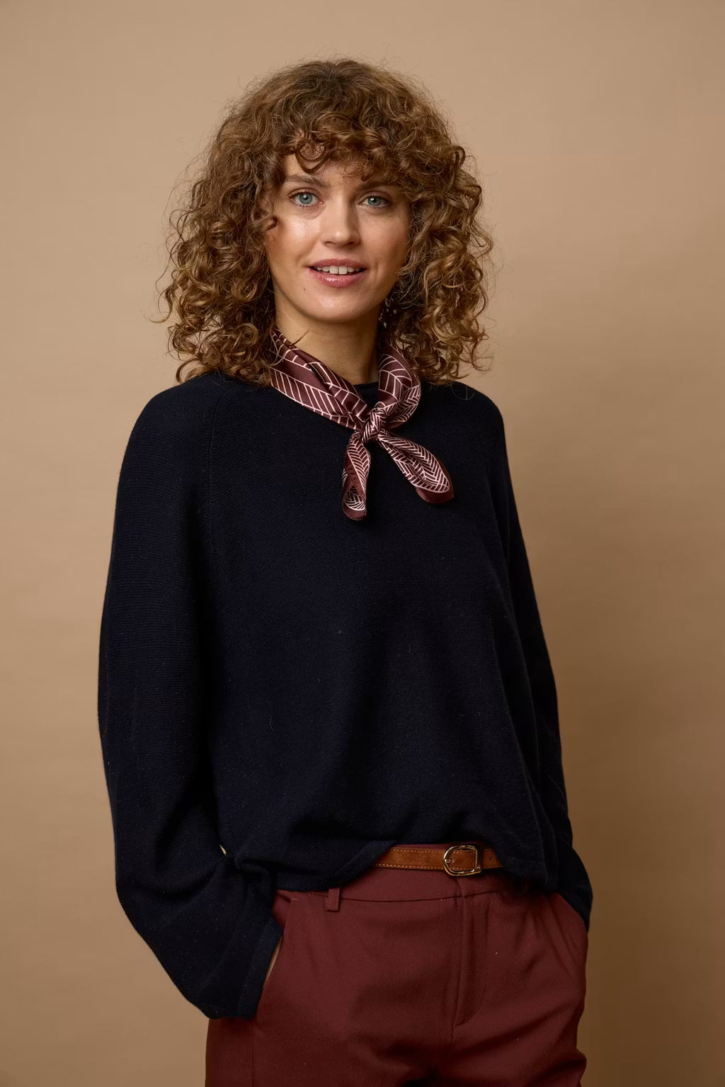 A woman with curly brown hair wearing a dark navy knit sweater, a burgundy and gold patterned neck scarf, and 
Rum Raisin / Red trousers, standing against a solid beige studio background.
