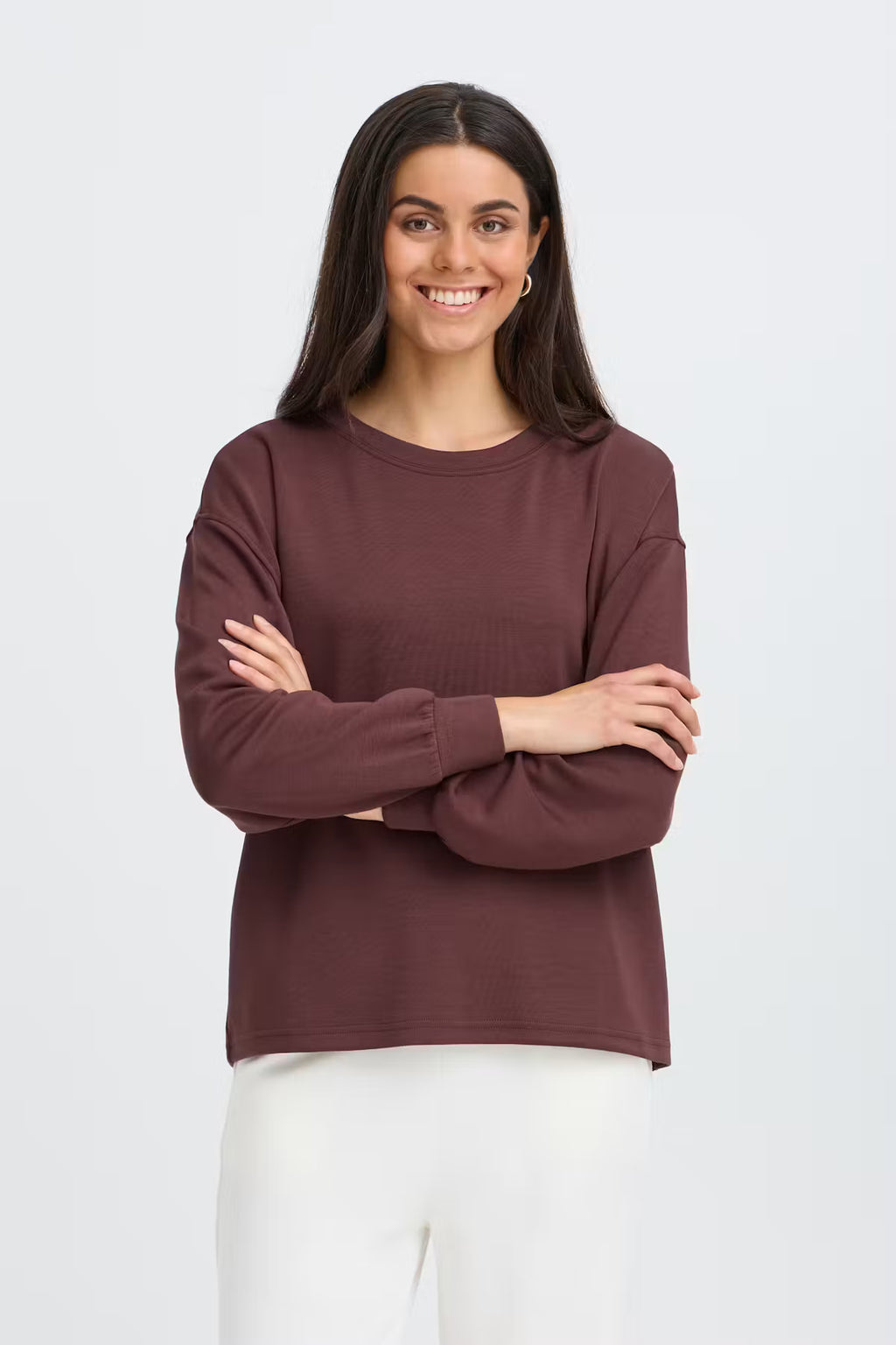 A woman with long dark hair stands against a light gray studio background wearing FRAURORA PU 7 Sweatshirt in Rum Raisin/Red   and white pants.