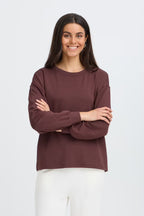 A woman with long dark hair stands against a light gray studio background wearing FRAURORA PU 7 Sweatshirt in Rum Raisin/Red   and white pants.