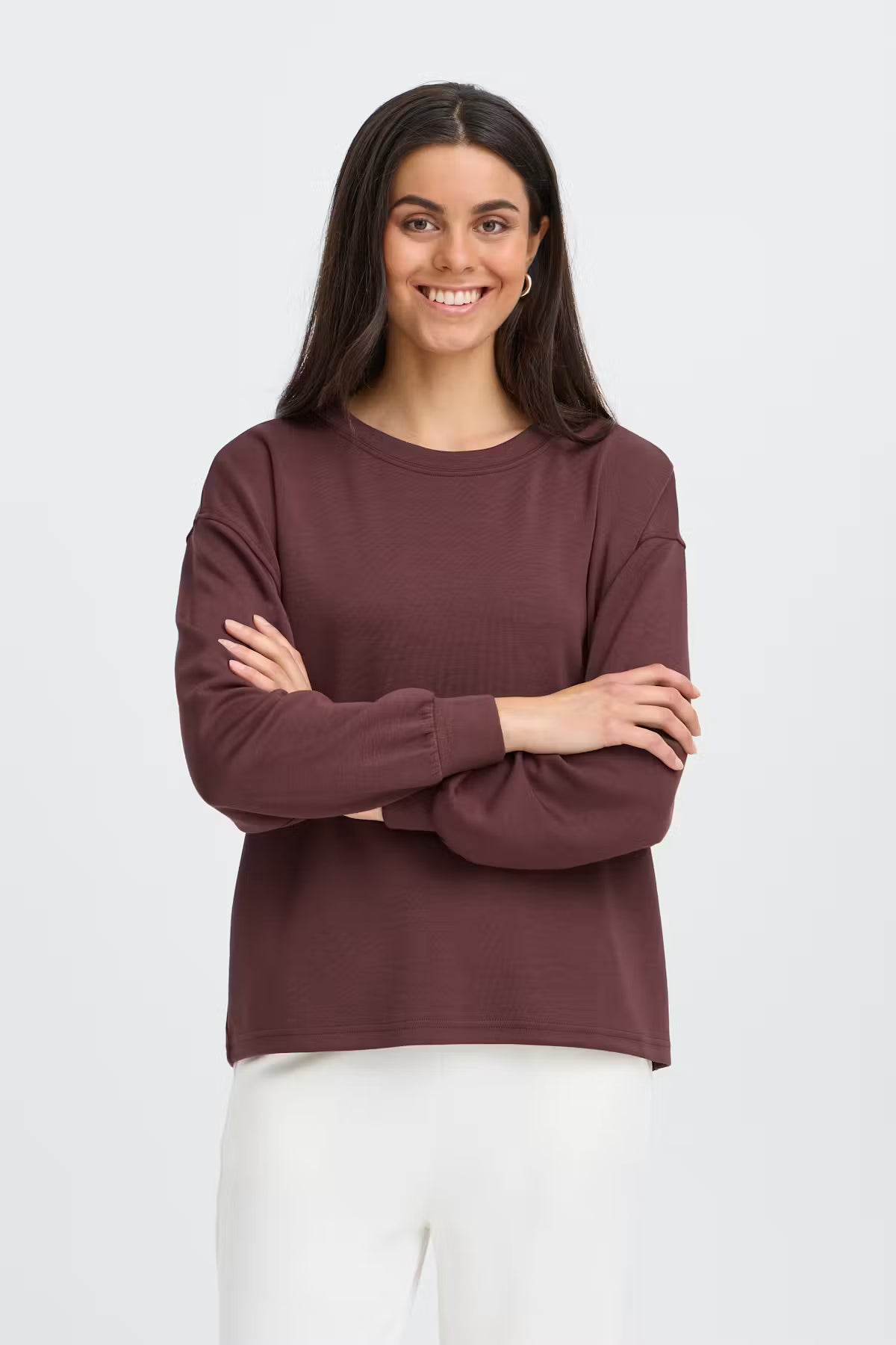 A woman with long dark hair stands against a light gray studio background wearing FRAURORA PU 7 Sweatshirt in Rum Raisin/Red   and white pants.