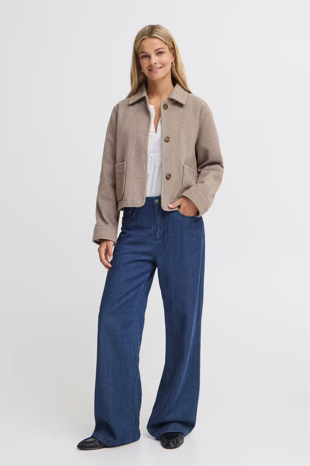 A woman modeling a cropped FREVILLA Outerwear in Rain-Drum Mix/Brown over a white top and high-waisted, wide-leg blue jeans, standing against a light gray studio background.