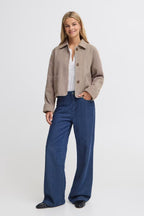 A woman modeling a cropped FREVILLA Outerwear in Rain-Drum Mix/Brown over a white top and high-waisted, wide-leg blue jeans, standing against a light gray studio background.