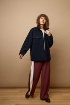 A model stands against a warm beige studio backdrop wearing a dark navy textured zip-up jacket over a white top, paired with wide-leg burgundy trousers featuring a white side stripe and dark brown loafers.