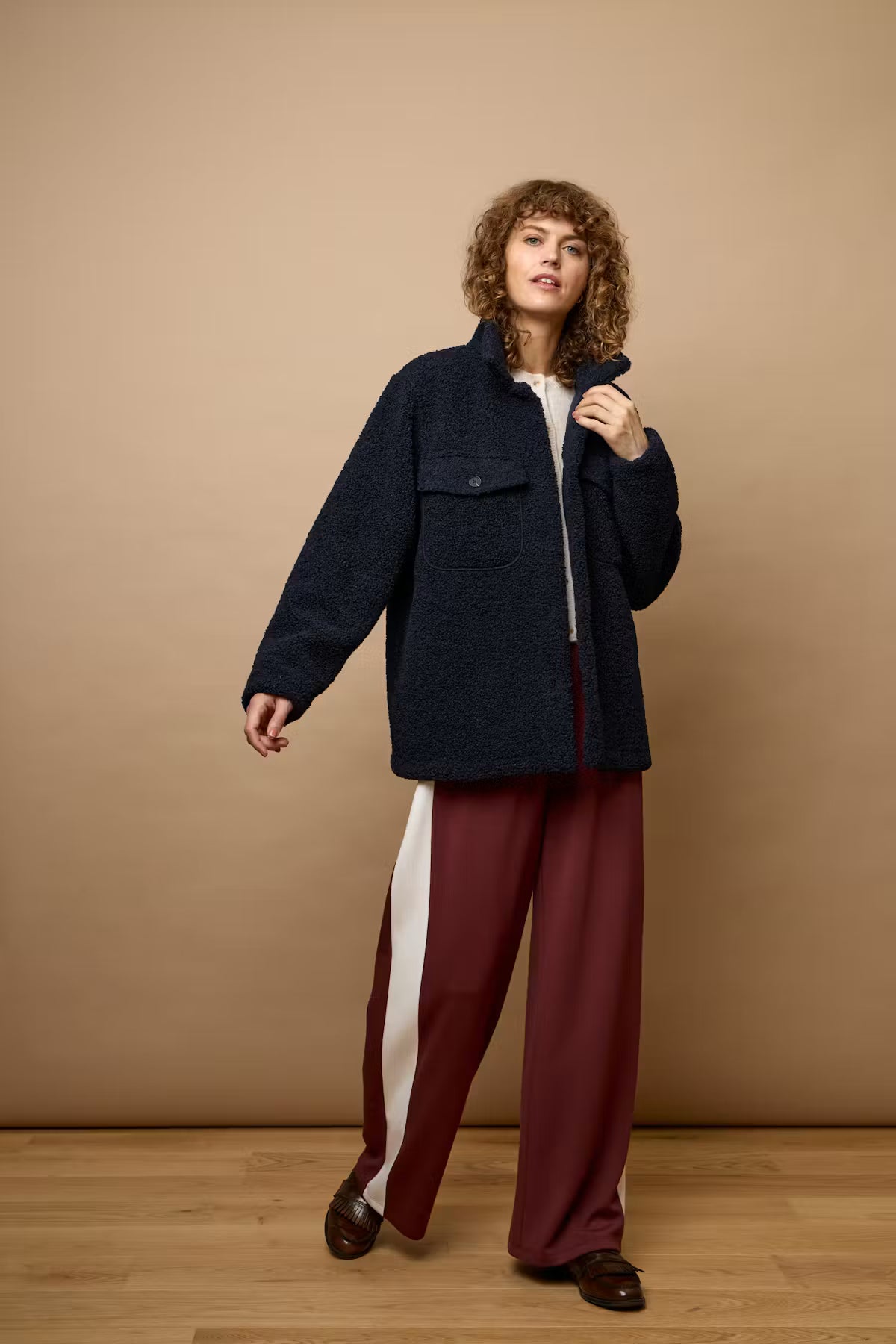 A model stands against a warm beige studio backdrop wearing a dark navy textured zip-up jacket over a white top, paired with wide-leg burgundy trousers featuring a white side stripe and dark brown loafers.