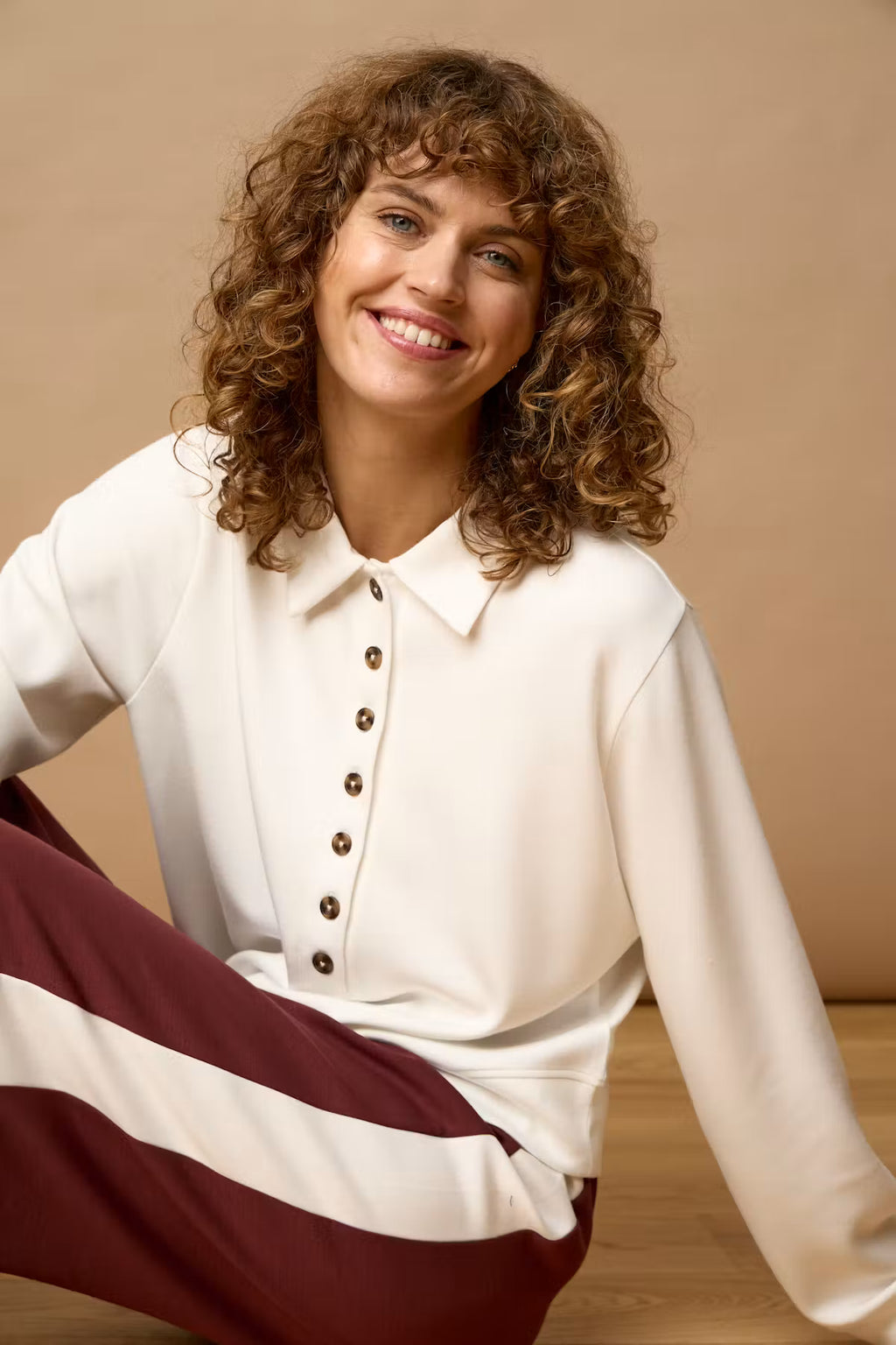 A smiling woman with curly brown hair sits on a wooden floor against a warm beige background, wearing a cream long-sleeve button-front collared top and burgundy-and-cream striped pants.