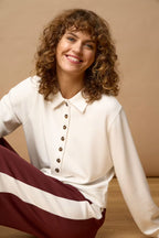 A smiling woman with curly brown hair sits on a wooden floor against a warm beige background, wearing a cream long-sleeve button-front collared top and burgundy-and-cream striped pants.
