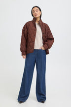 A model stands against a light gray studio background wearing a FRAGATE Outerwear in Rum Raisin/Red over a cream top and blue wide-leg jeans.
