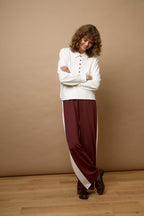 A woman with curly brown hair stands in a studio wearing a white long-sleeve henley top and wide-leg burgundy pants with white side panels, posed against a solid tan backdrop and light wood floor.