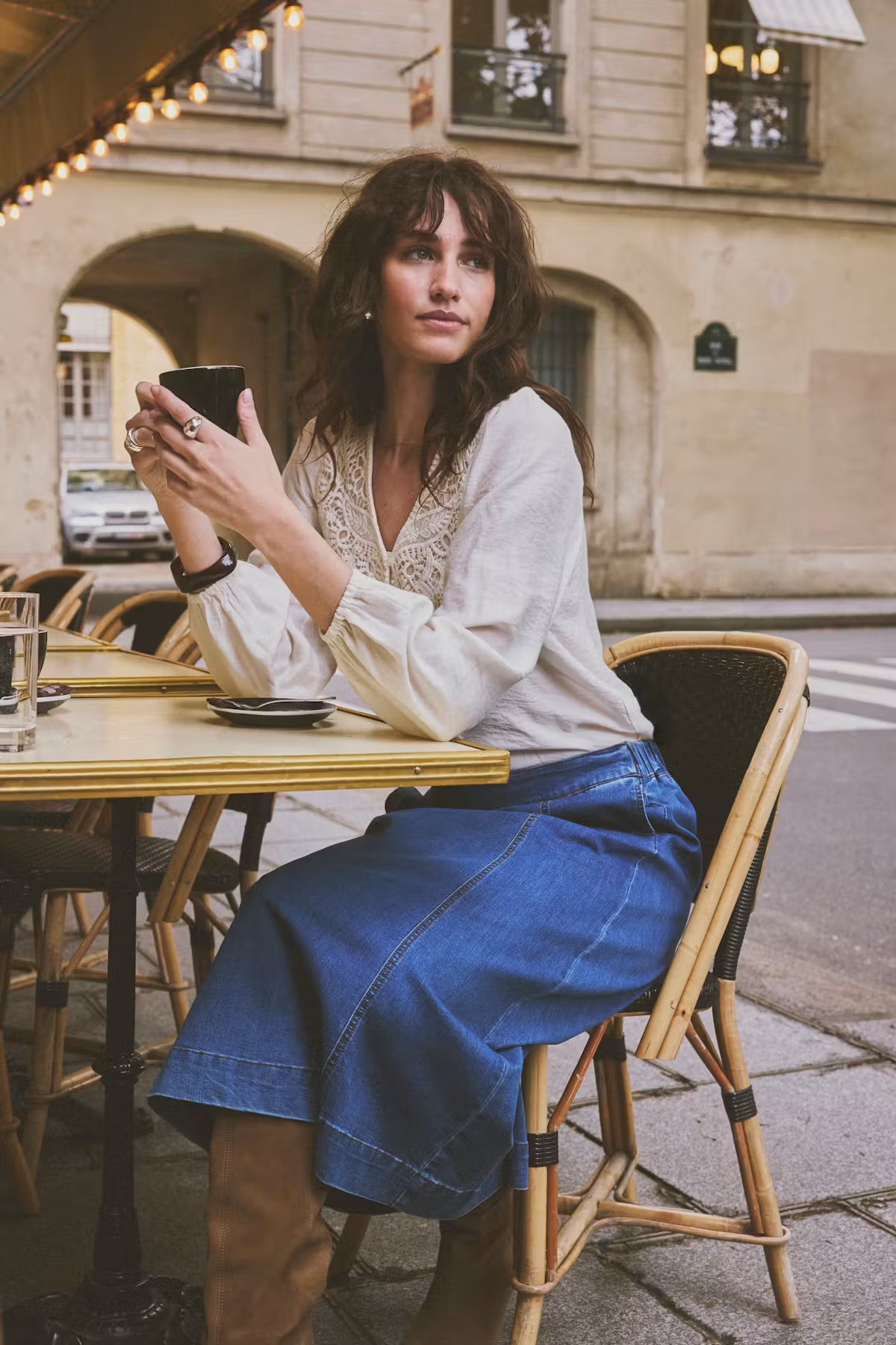 A woman sits at an outdoor cafe table wearing a Whisper White blouse and a blue denim skirt, holding a black coffee cup. The background shows a European-style street with stone buildings and arches.