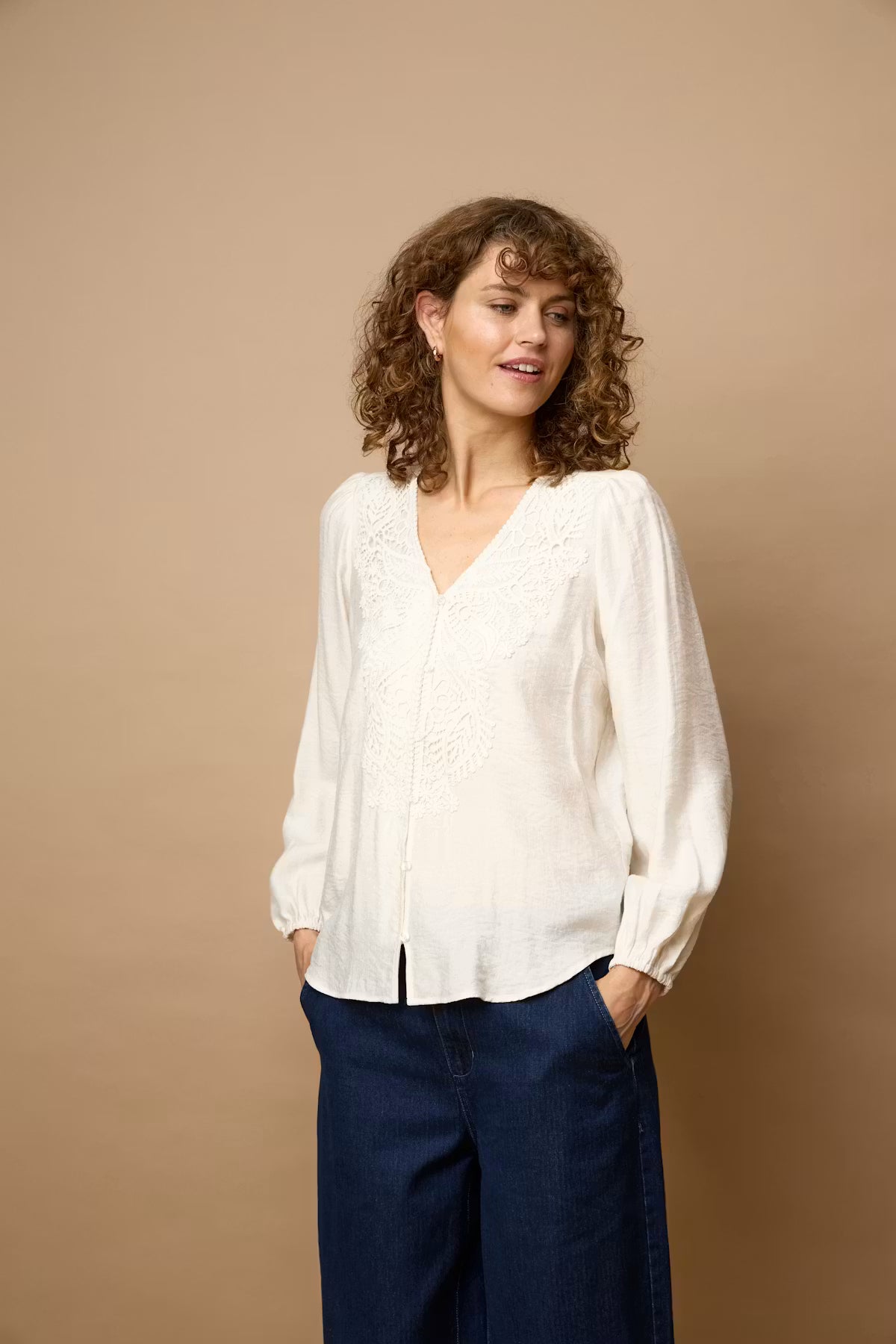 A woman with curly brown hair wearing a Whisper White long-sleeve blouse with delicate embroidery and dark blue wide-leg trousers, standing against a solid beige studio background.